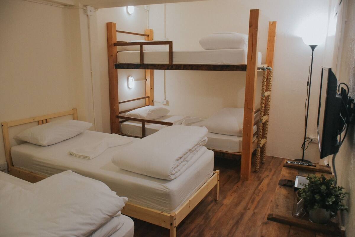 A bunk bed room with wooden frames, white bedding, a bedside table, and a floor lamp.