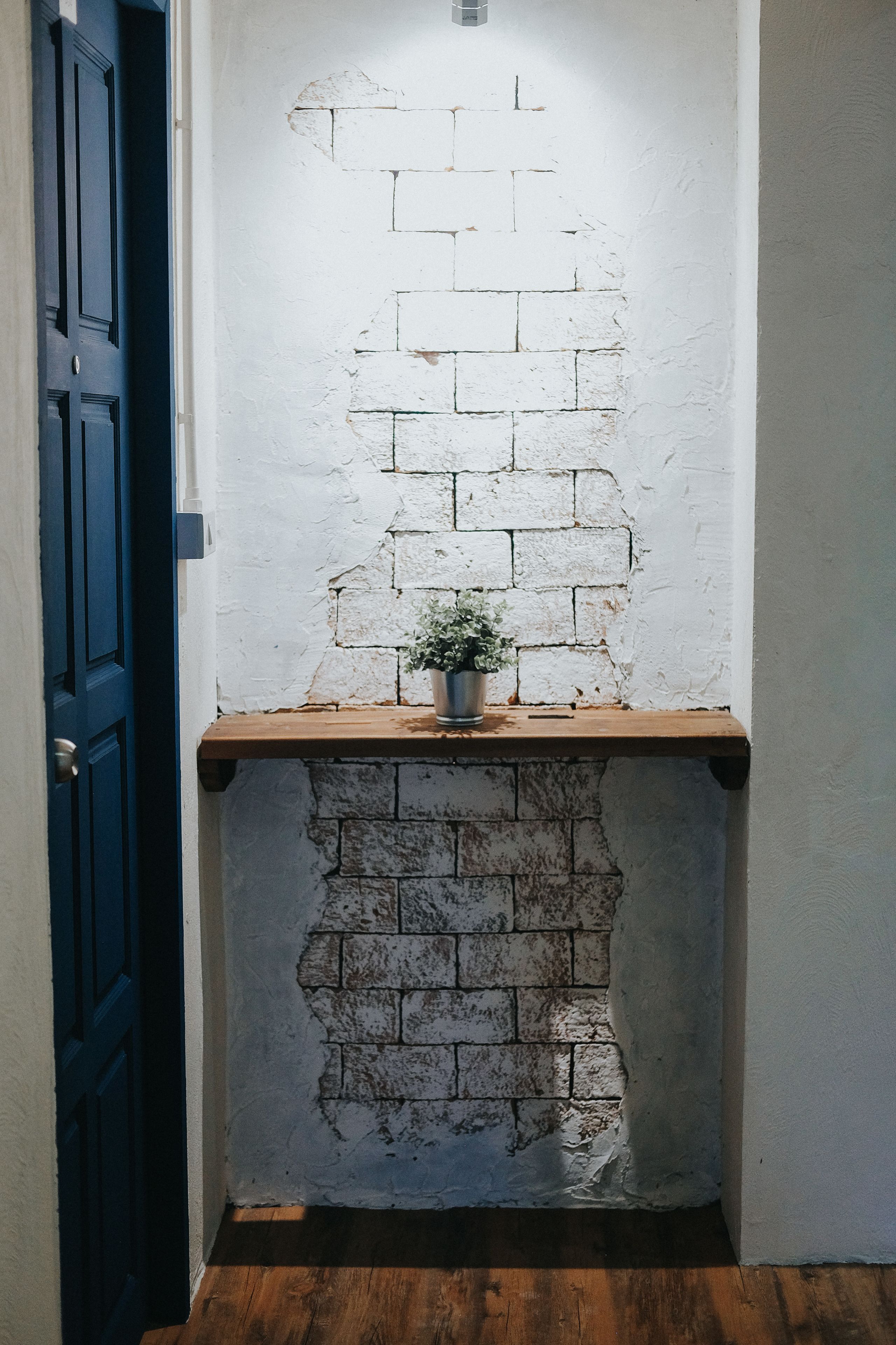 A narrow hallway with a white brick wall, a wooden shelf with a potted plant, and a blue door.