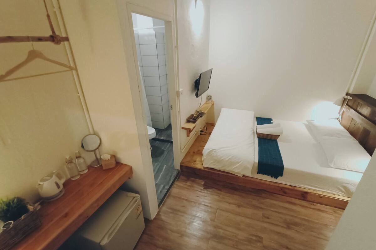A small, compact bedroom with a wooden bed, a bedside table, a TV, and a small window.