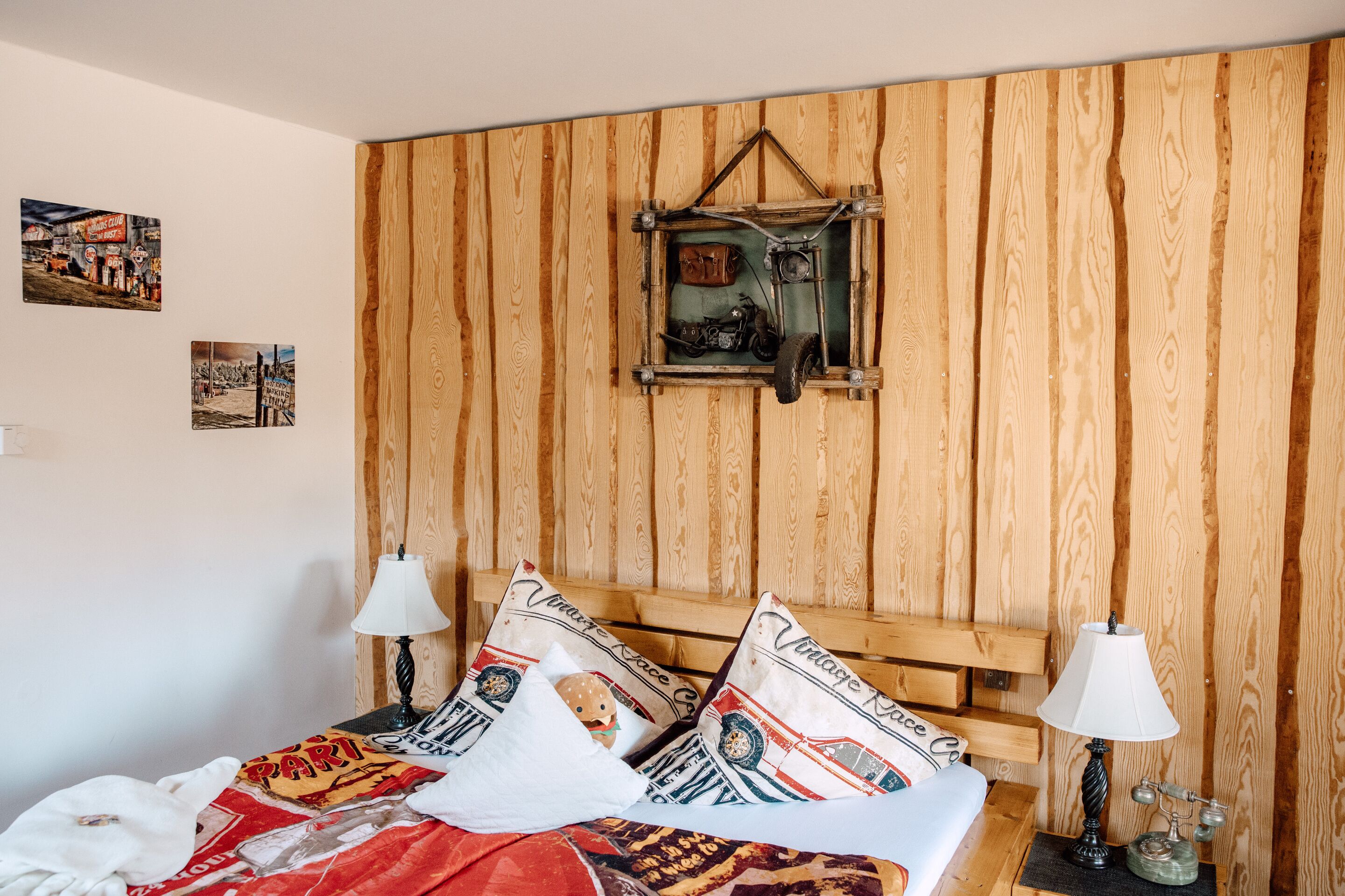 A bedroom with a wooden headboard, two bedside lamps, a bed with patterned bedding, and a vintage-style wall hanging.