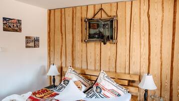 A bedroom with a wooden headboard, two bedside lamps, a bed with patterned bedding, and a vintage-style wall hanging.