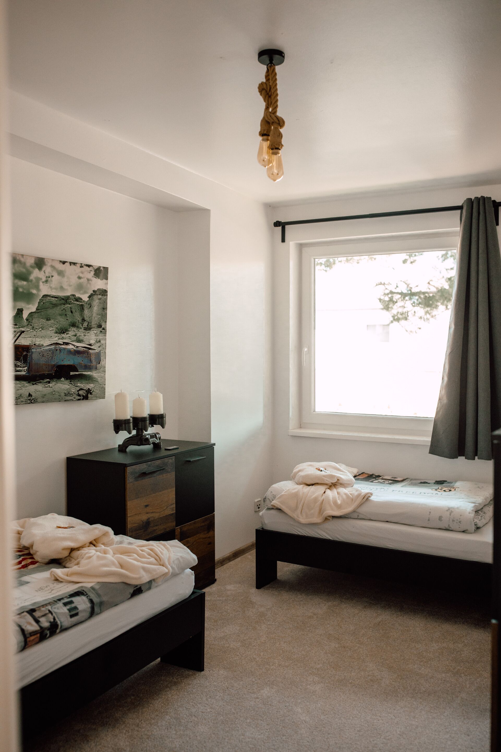 A bedroom with two beds, a dresser, a window with curtains, and a wall-mounted light fixture.