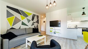 Una sala moderna con un sofá gris, una mesa de centro blanca y un sillón negro. Se ve un área de cocina con armarios blancos y una encimera negra al fondo.