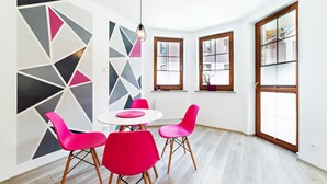 Un comedor moderno con una mesa blanca, cuatro sillas rosadas, arte mural geométrico y ventanas amplias con persianas.
