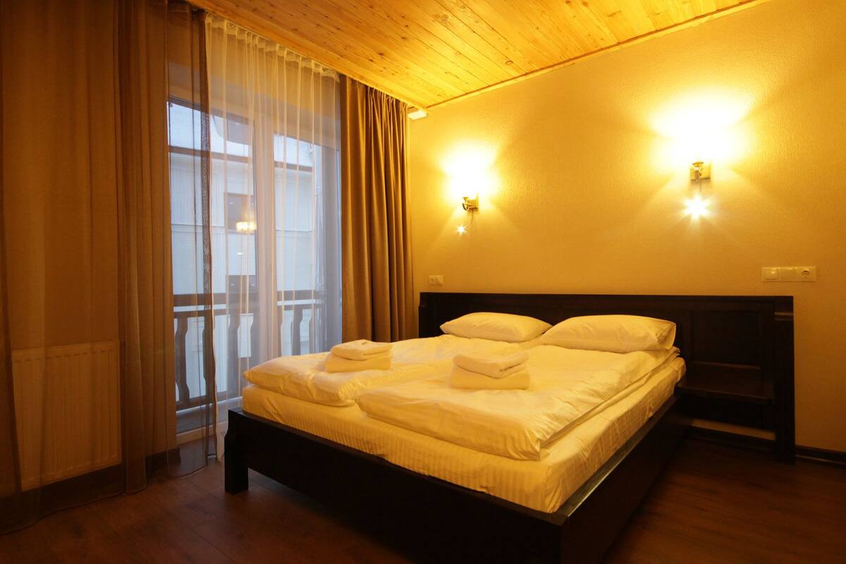 A hotel room with a large bed, wooden headboard, two wall sconces, a window with sheer curtains, and a balcony view.