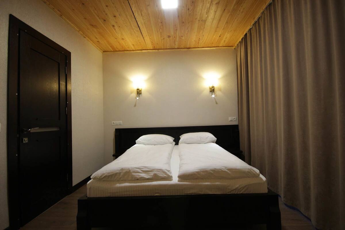 A double bed with two pillows, a wooden ceiling, and two wall-mounted lights.