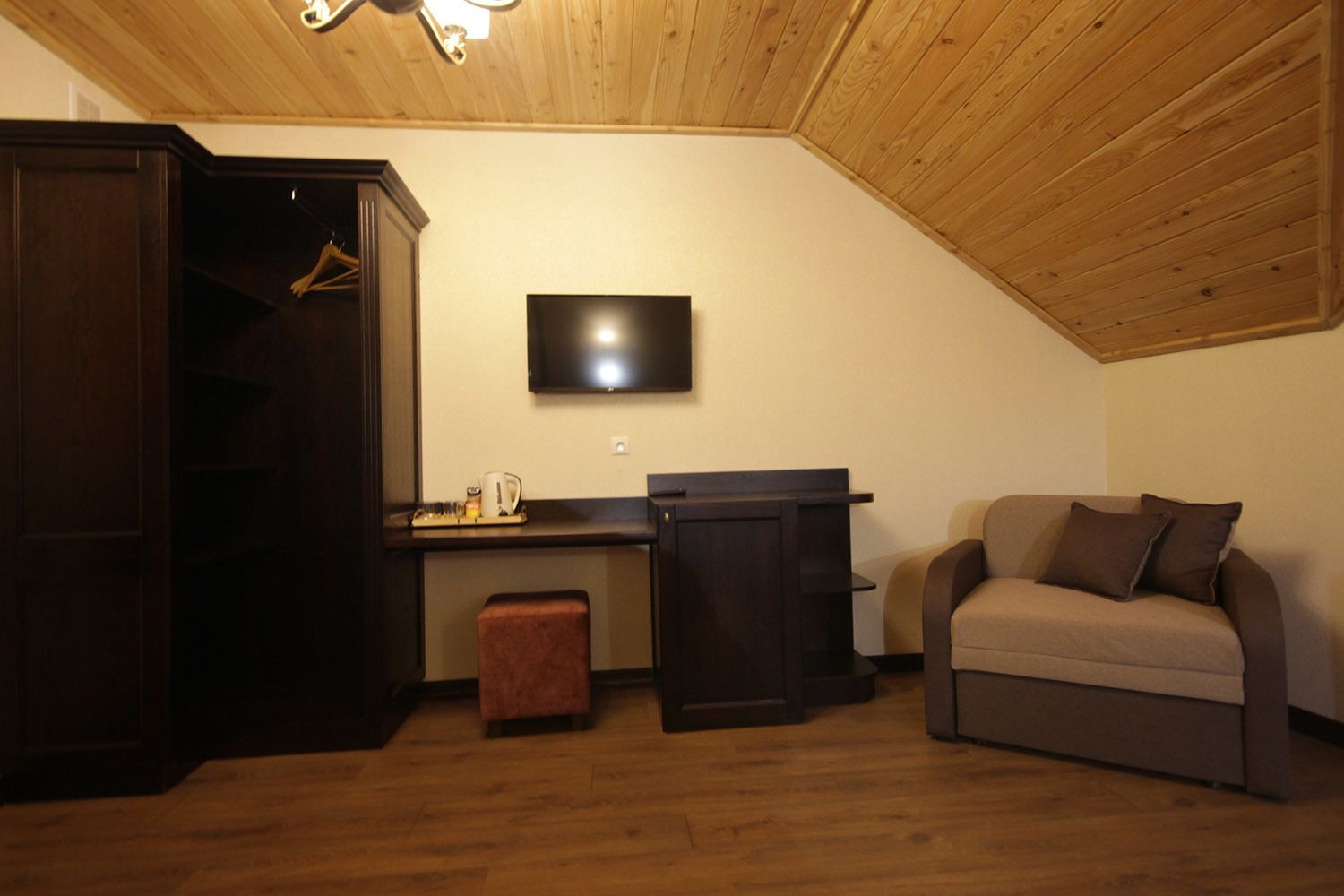 A cozy room with a wooden ceiling, a TV mounted on the wall, a desk with a chair, a sofa, and a wardrobe.