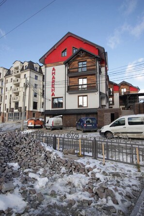 A multi-story building with a red and white facade, a visible sign with the name 'AMARINA,' and several parked vehicles in front. - Amarena (Bukovel)