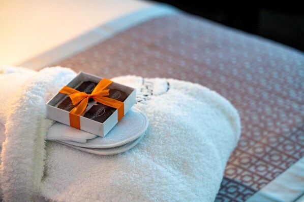 A neatly folded white towel with a small box of chocolates placed on top, tied with an orange ribbon. - Nash Pratik Hotel (Meyrin)