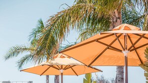 Een dakterras met witte ligbedden en oranje parasols, omgeven door tropische planten en palmbomen.