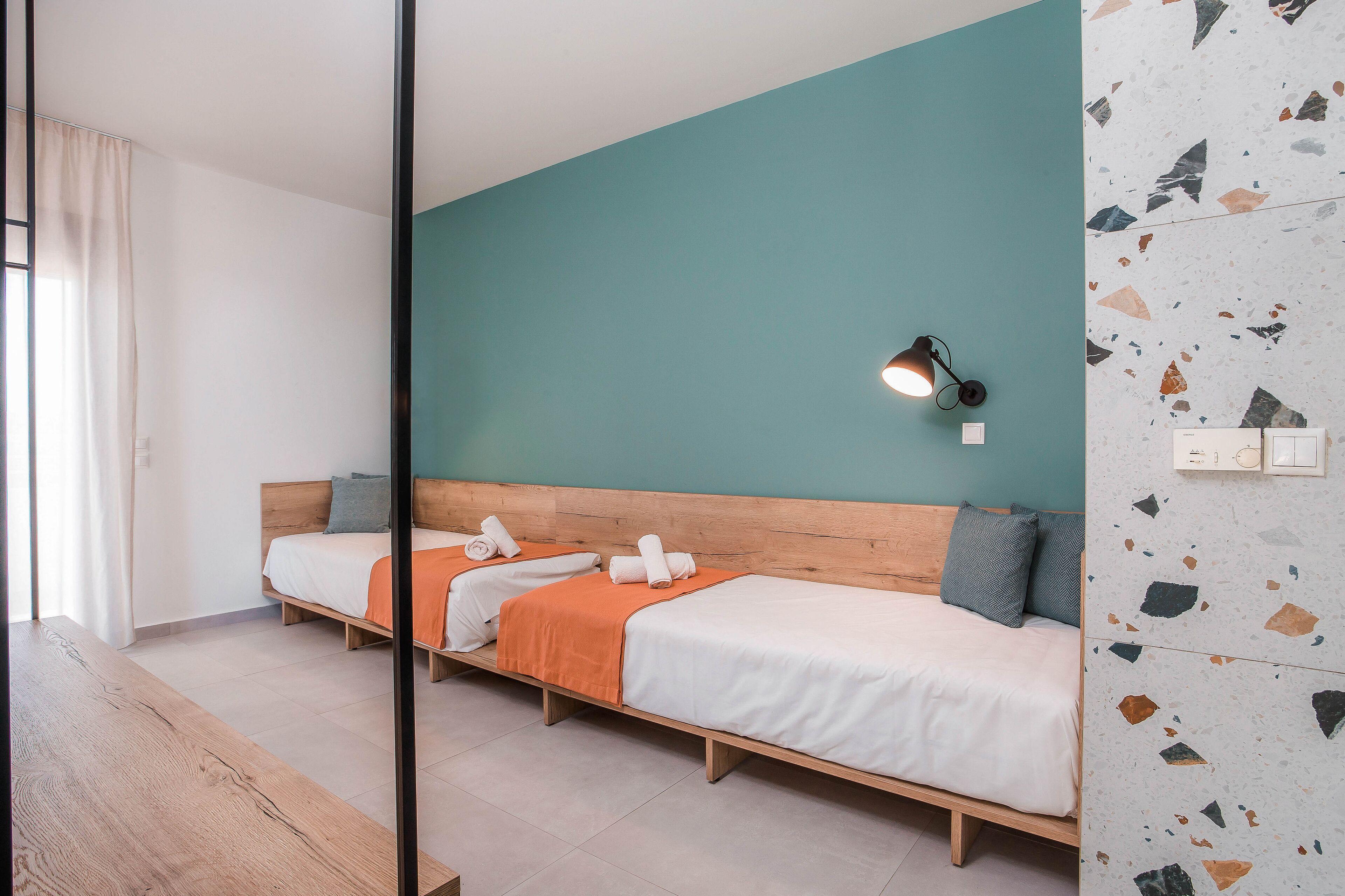 Ett hotellrum med två separata sängar, en träbänk, en vägg i en turkos nyans som accentfärg och en vägg med terrazzomönster.