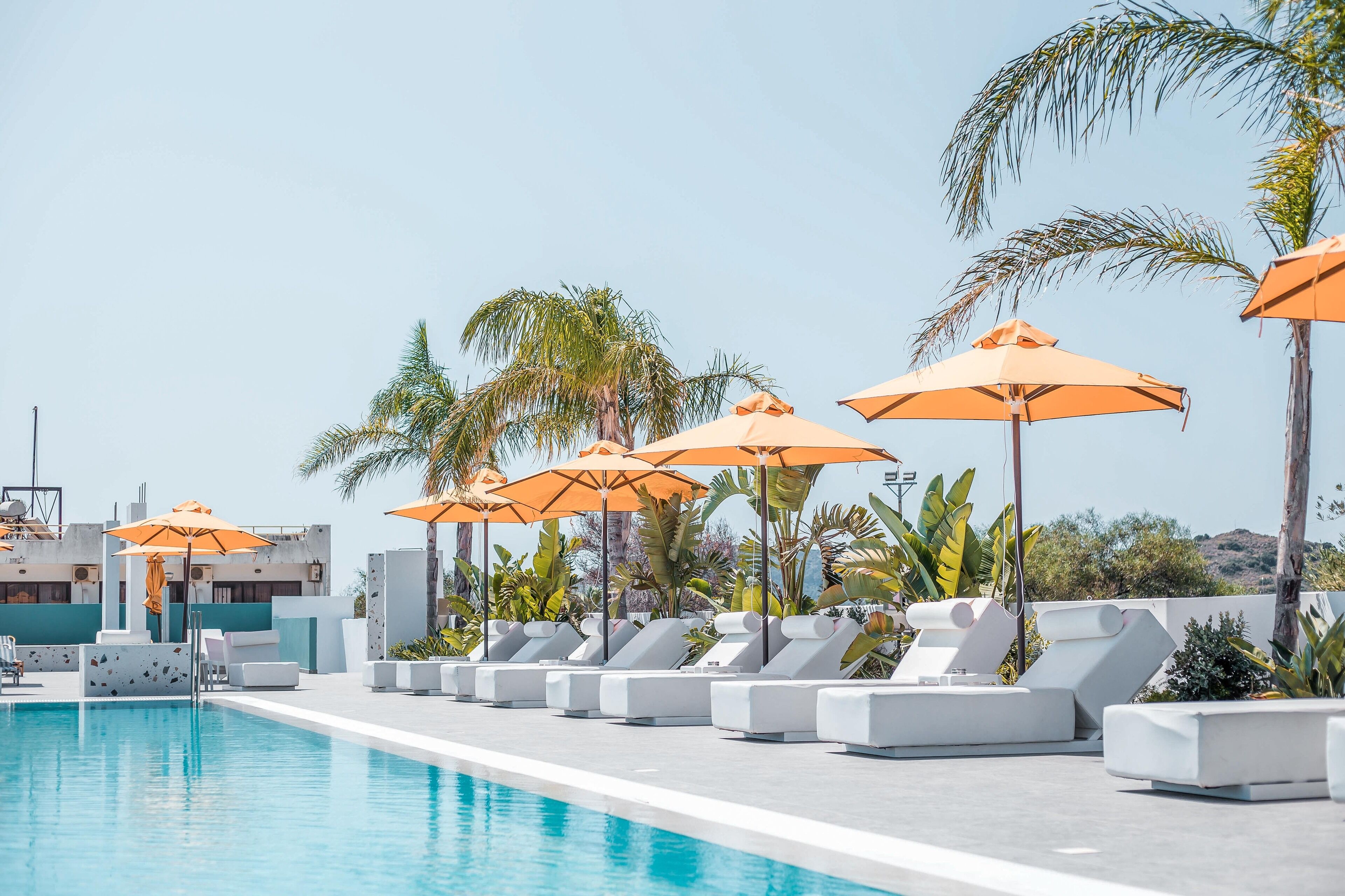 A poolside area with lounge chairs and orange umbrellas, surrounded by palm trees and a clear sky.