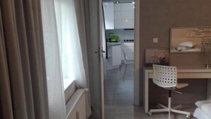 A hotel room with a bed, a desk with a chair, a window with curtains, and a view of the kitchen. - EverySky Karpacz 3 Maja 66 (Karpacz)