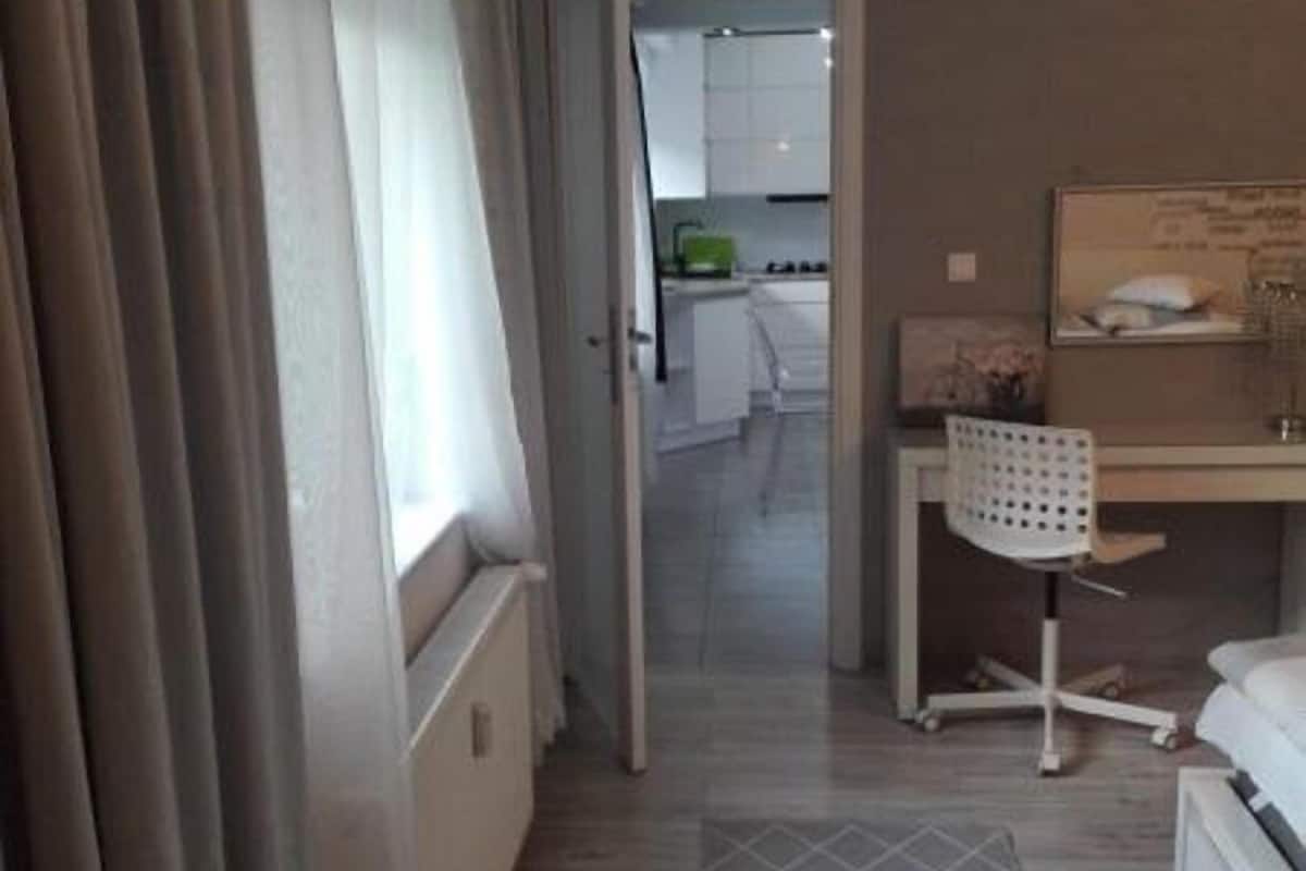 Quarto de hotel com uma cama, uma escrivaninha com cadeira, uma janela com cortinas e vista para a cozinha.