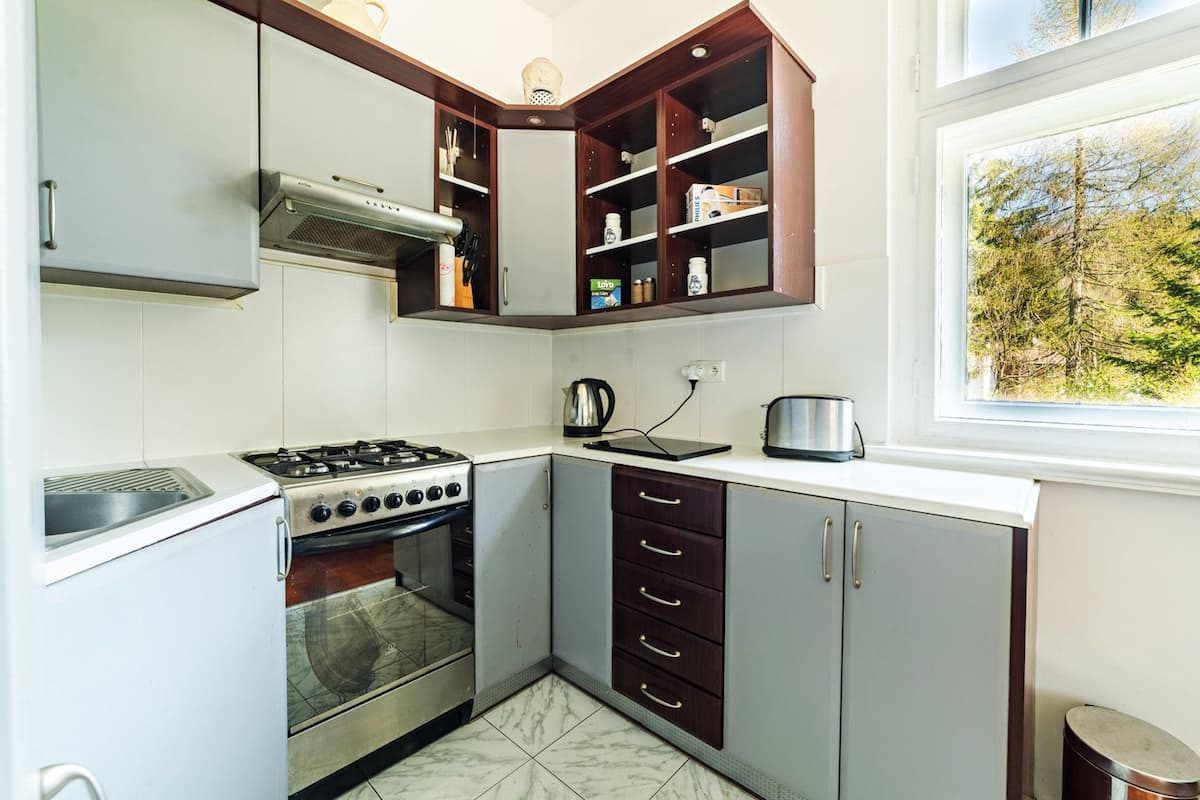 Cozinha compacta com fogão a gás, forno e micro-ondas embutido. Possui uma janela com vista para árvores, armários de madeira e painel de azulejos.