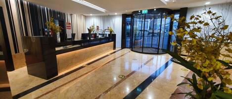 A modern hotel lobby with a reception desk, a glass elevator, and a marble floor.