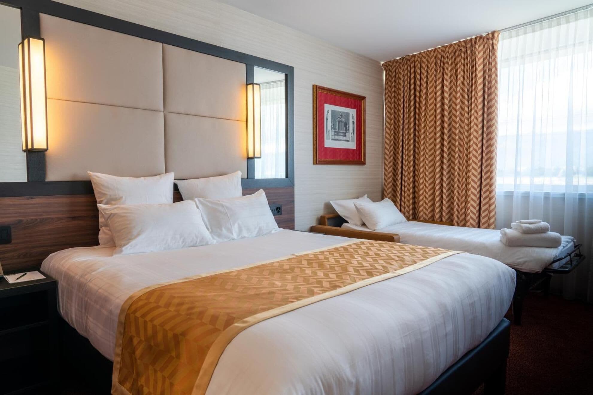 Una habitación de hotel con dos camas, una lámpara grande de pared, un cuadro enmarcado y una ventana con cortinas.