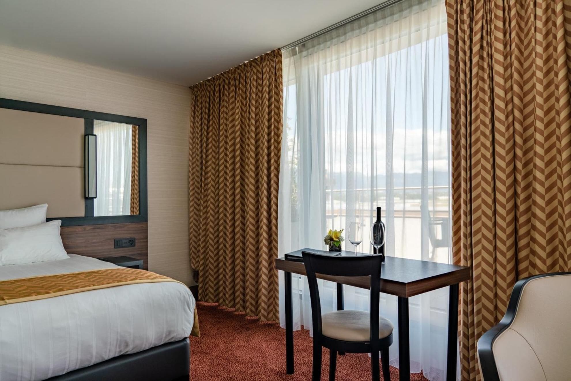 Een hotelkamer met een bed, een bureau met stoel, een spiegel en een raam met gordijnen.