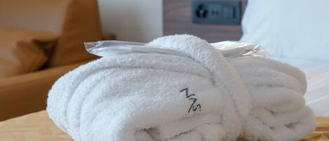 A stack of white towels on a patterned bedspread in a hotel room with a brown leather sofa and a white pillow in the background.