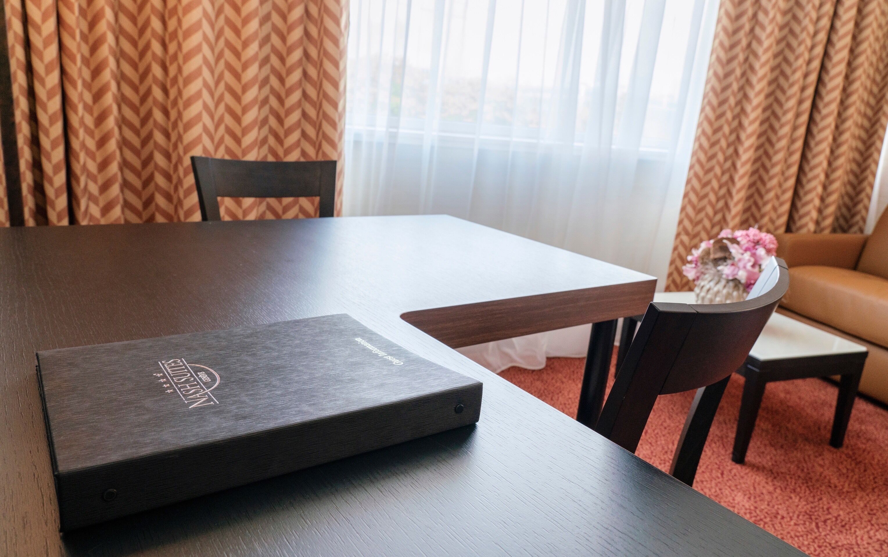 A hotel room with a wooden table, a black folder, and a vase with flowers on a side table.