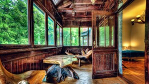 A rustic cabin interior with wooden walls and floors, large windows offering views of a forest, and a central table with antler stools. - EverySky Piastowska1 Karpacz (Karpacz)