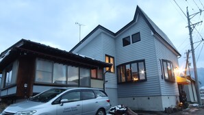Een gebouw met twee verdiepingen, een schuin dak, een auto ervoor en een stapel hout naast het gebouw.