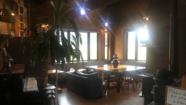 Een gezellige ruimte met een houten tafel, een bank en een grote plant. Er zijn ramen waardoor natuurlijk licht binnenkomt, en een bord met tekst aan de muur.