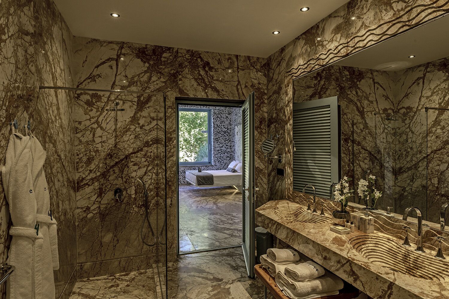 Ein Badezimmer mit Marmorwänden, einem großen Spiegel, einer Duschkabine aus Glas und Blick auf ein Schlafzimmer durch eine offene Tür.
