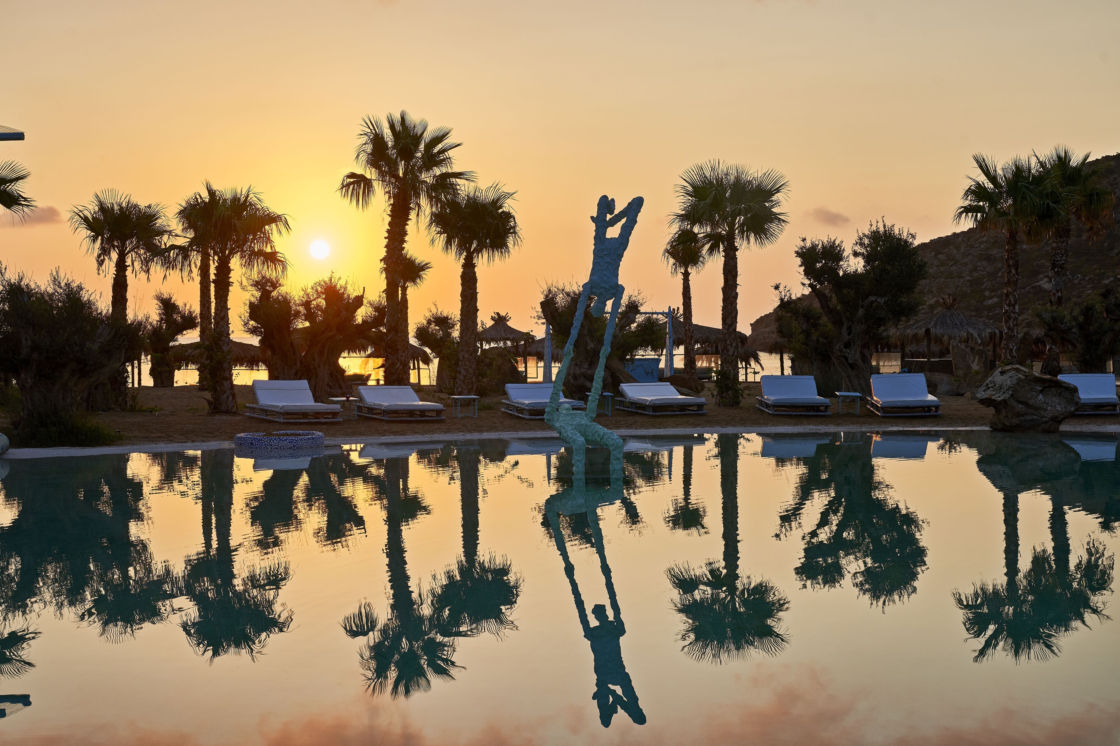 Une piscine agrémentée d’une sculpture, de palmiers et de chaises longues, le tout baigné par les couleurs du coucher de soleil.
