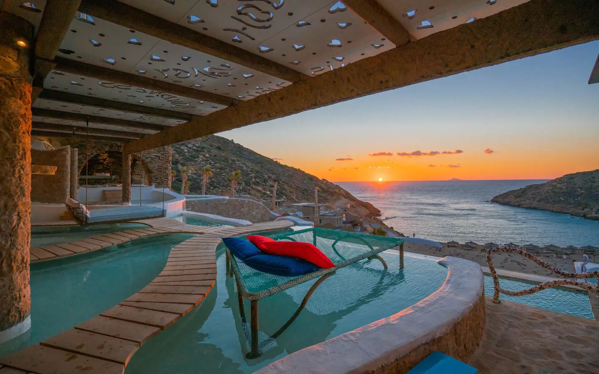 A pool area with a view of the sunset over the sea, stone pillars, and a lounge chair with cushions.