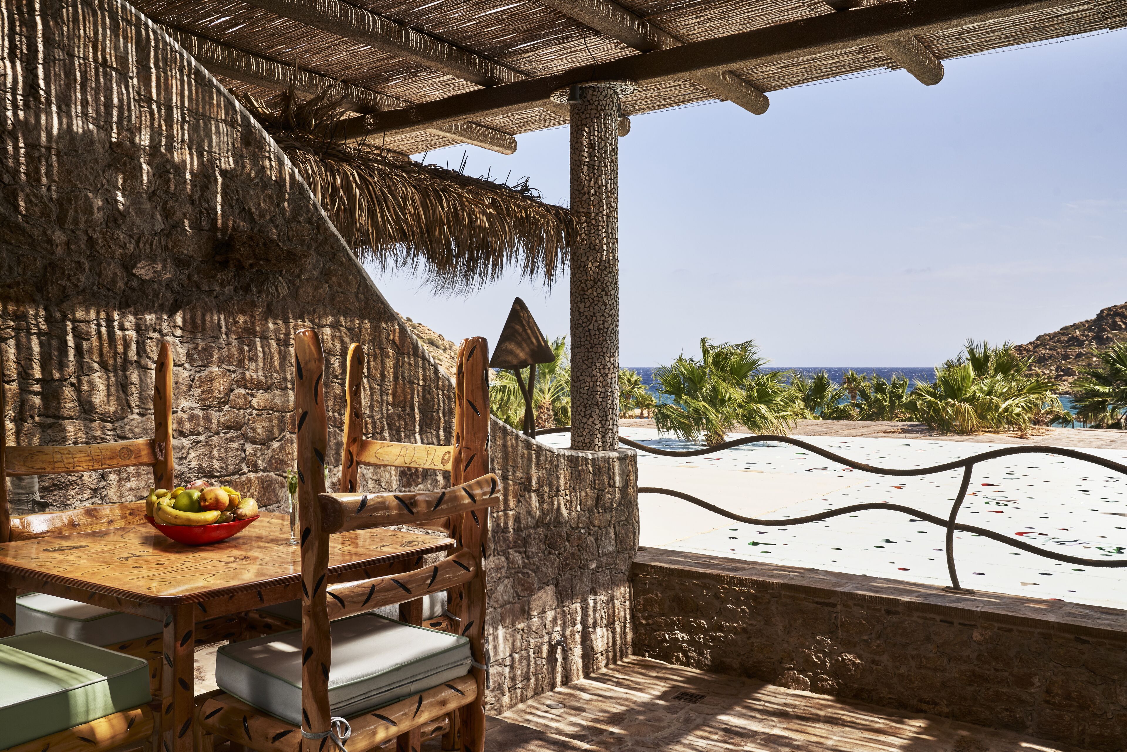 Ein Holztisch mit Obstschale, eine Steinwand und Blick auf Strand und Meer.