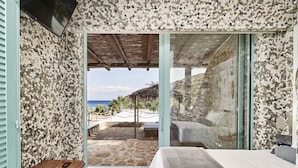 A bedroom with a bed, a small table, and a view of a beach and ocean through a glass door. - Calilo (Ios)