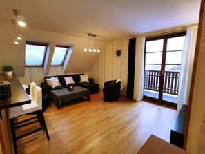 A modern living room with a sofa, coffee table, and a dining area with a table and chairs. There is a balcony with a railing and a view of the outside.