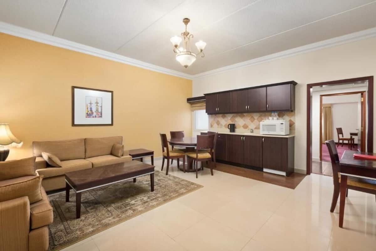 A living room with a sofa, coffee table, and a dining area with chairs and a table. There is a kitchen area with cabinets and a microwave.