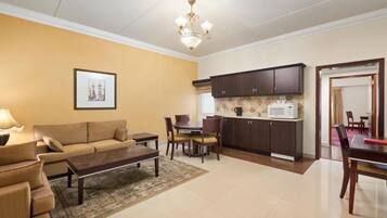 A living room with a sofa, coffee table, and a dining area with chairs and a table. There is a kitchen area with cabinets and a microwave.