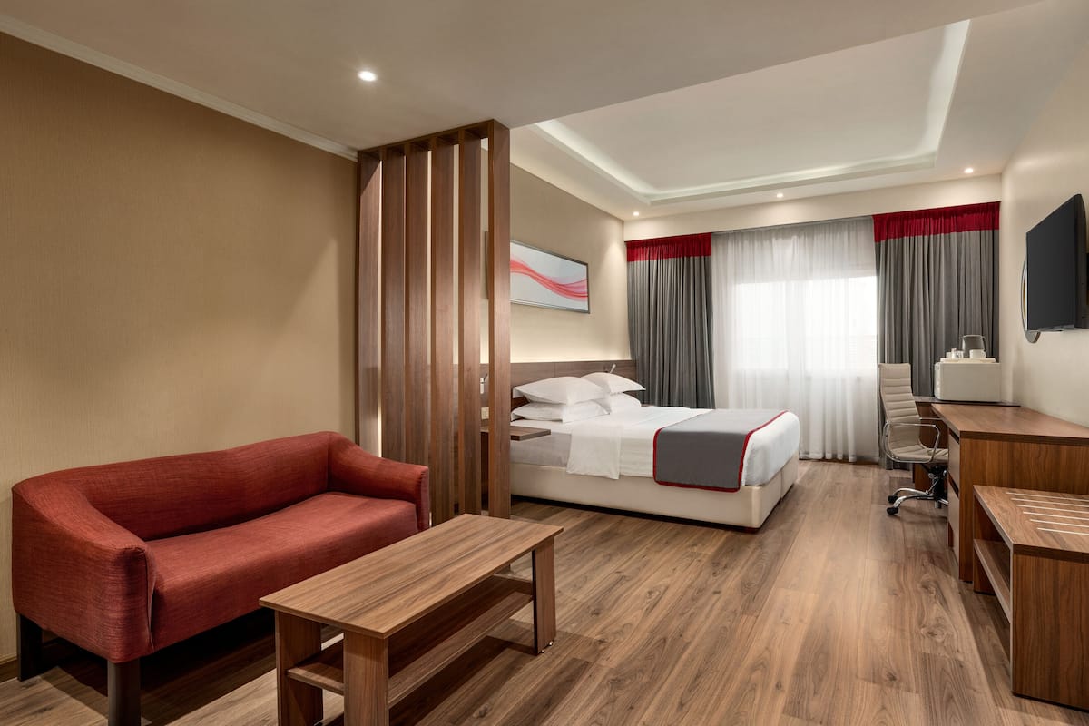 A hotel room with a red sofa, a wooden desk, a bed with white bedding, a TV, and a window with grey curtains.