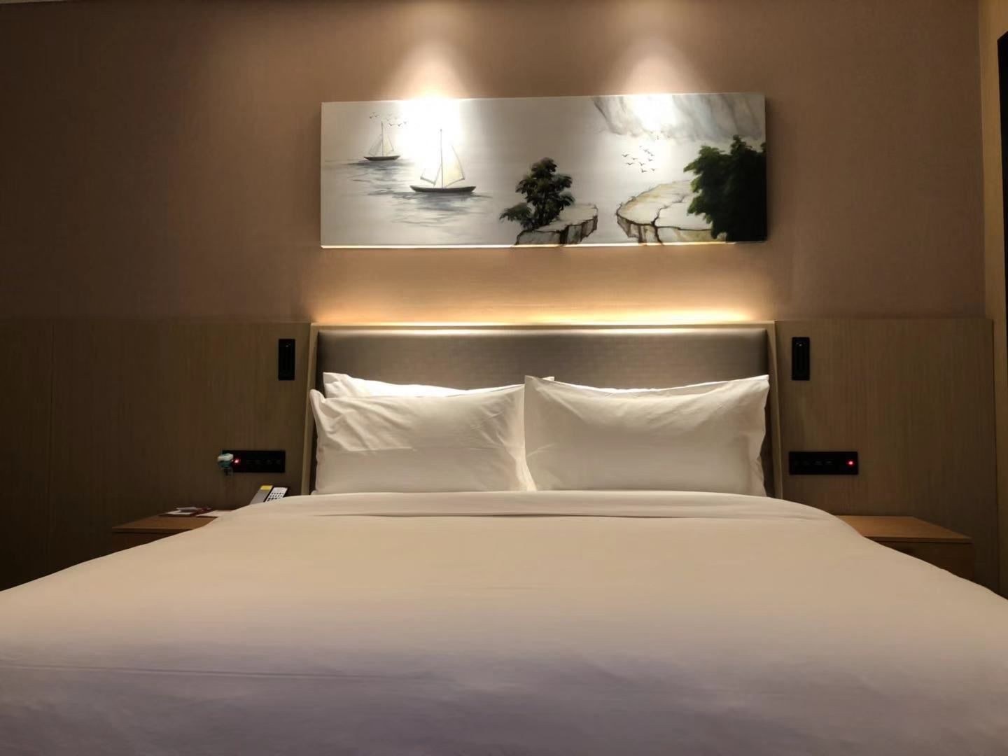 Hotellihuone, jossa on suuri sänky, kaksi yöpöytää, yölamppu ja seinälle ripustettu taulu, jossa on kuvattuna purjeveneitä ja puita.