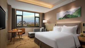 A hotel room with a large bed, a desk with a chair, a small sofa, a large window with a city view, and a painting on the wall. - Ramada Wyndham Zhengzhou Xinzheng (Zhengzhou)