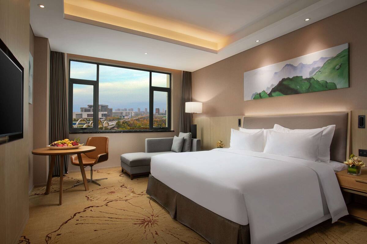 Ein Hotelzimmer mit einem großen Bett, einem Schreibtisch mit Stuhl, einer kleinen Sitzecke und einem großen Fenster mit Blick auf die Stadt. An der Wand hängt ein Gemälde.