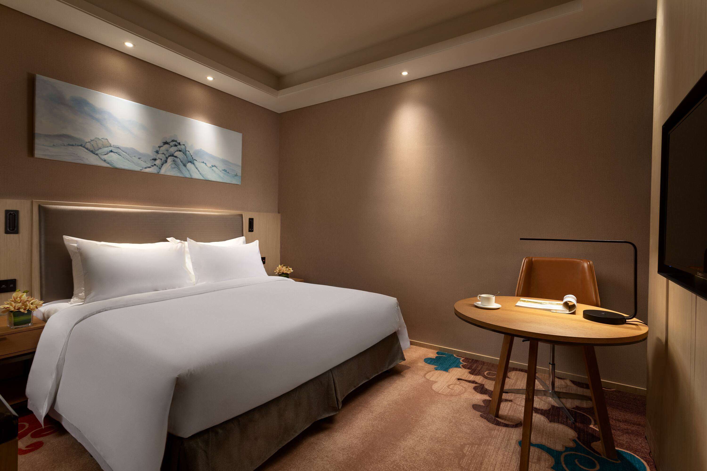 A hotel room with a large bed, a small table, a chair, a painting on the wall, and a TV mounted on the wall.