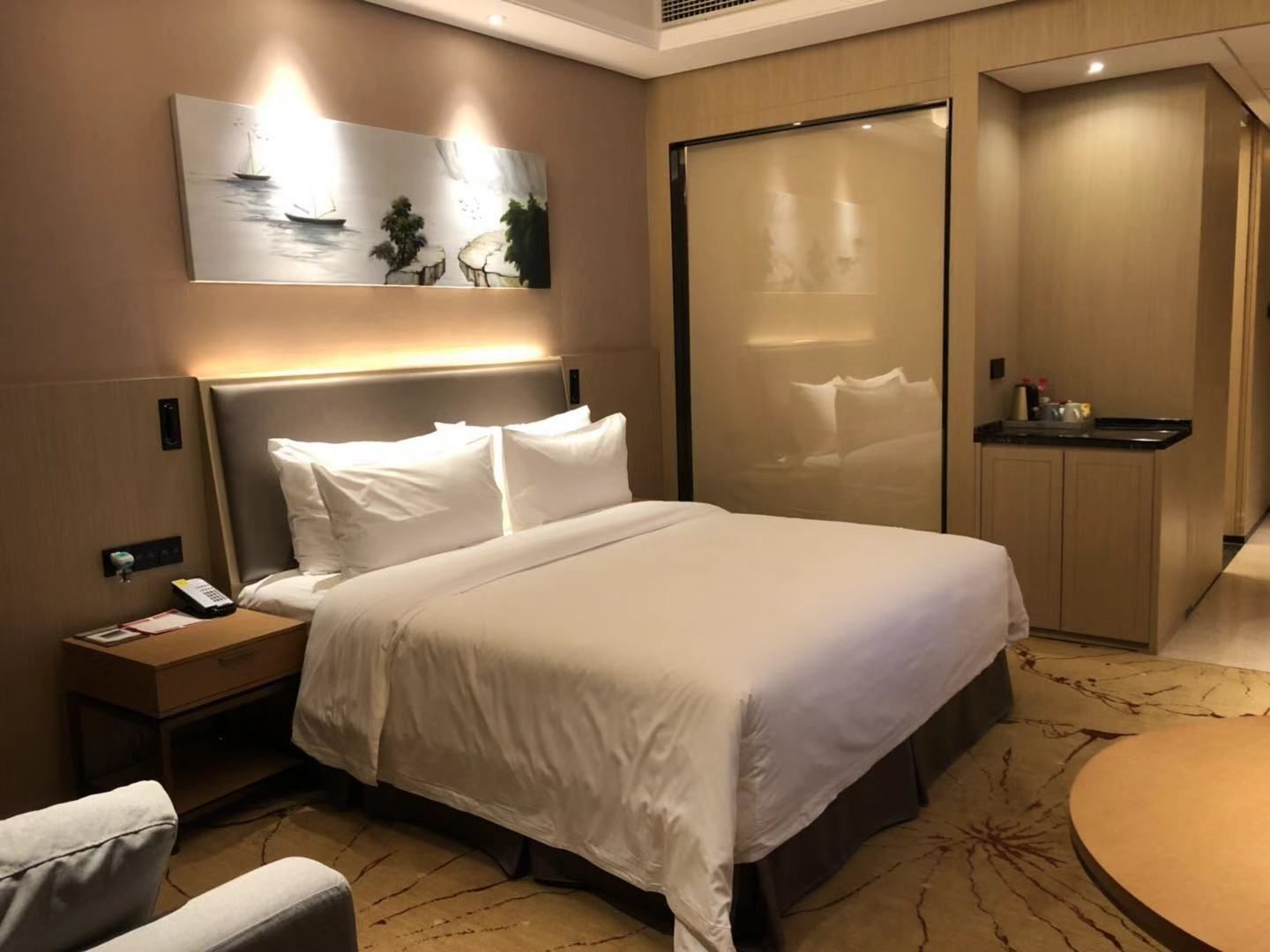 Une chambre d’hôtel avec un grand lit, une table de chevet avec un téléphone, un petit bureau, une chaise, une table ronde et un tableau au mur.