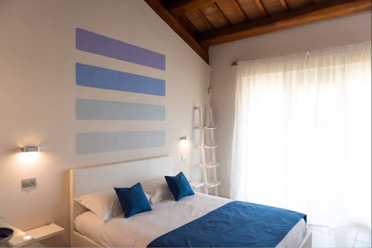 A bedroom with a bed, a nightstand, a bedside table, a wall-mounted shelf, and a window with curtains.