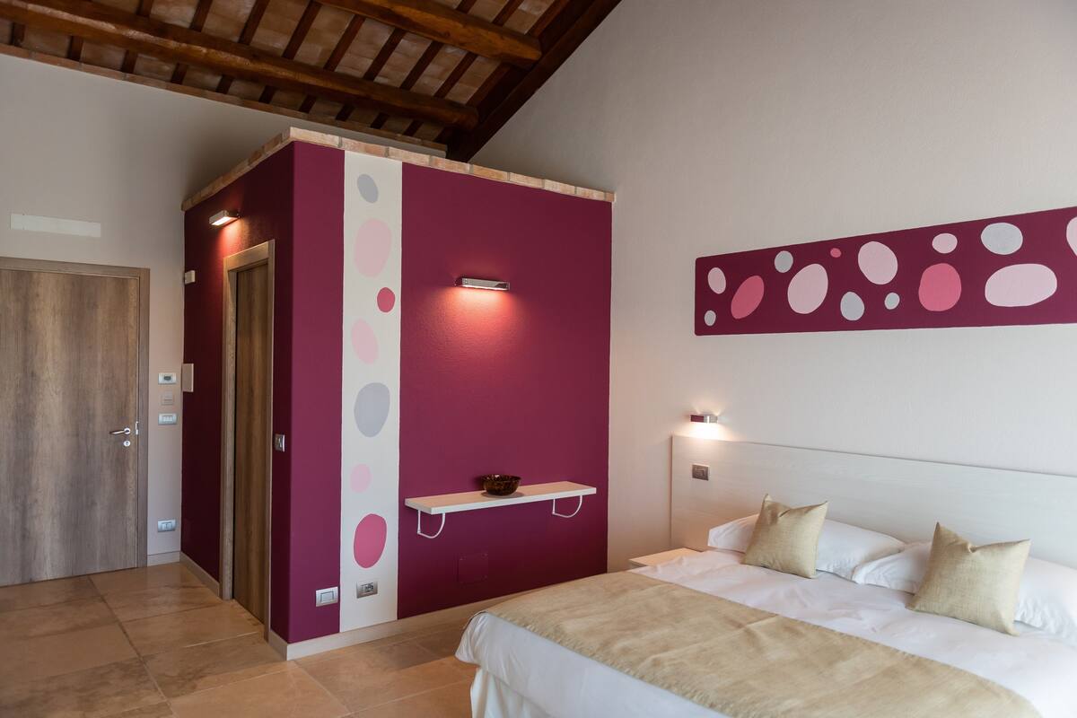 A bedroom with a bed, a nightstand, a wall-mounted shelf, a door, and a ceiling with exposed beams.