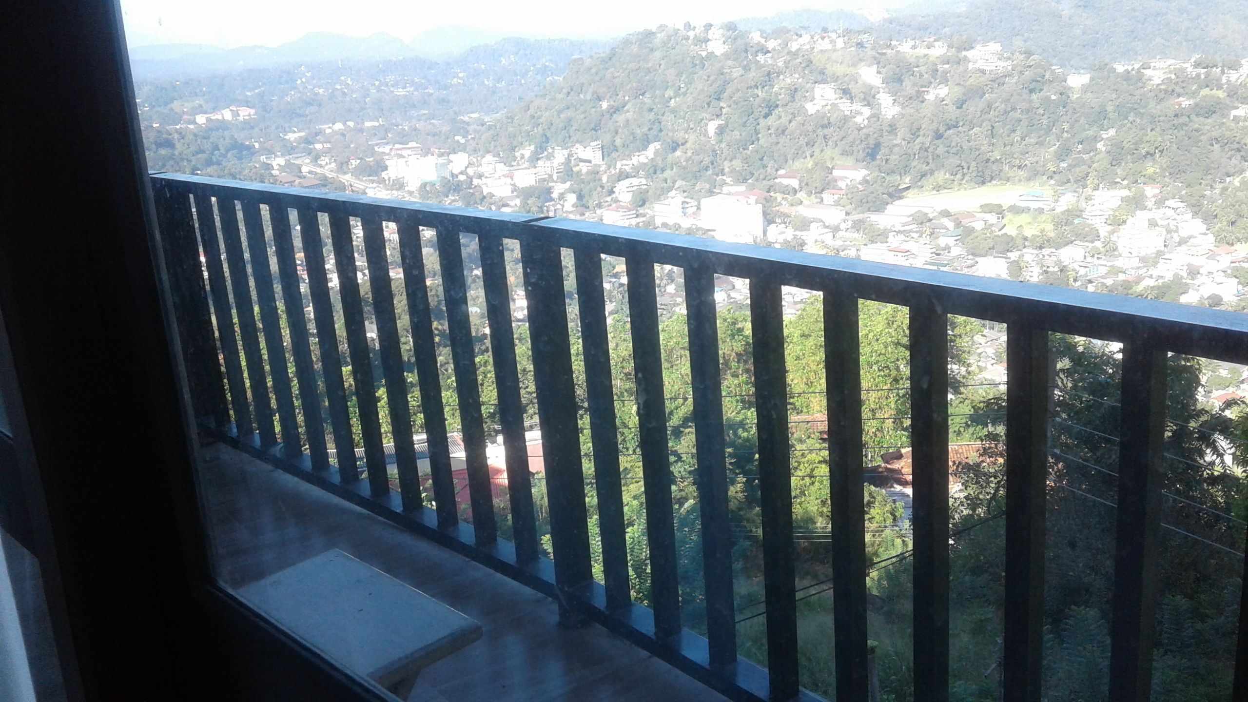 Un balcon doté d’une rambarde en métal, donnant sur une ville avec ses bâtiments et sa végétation.