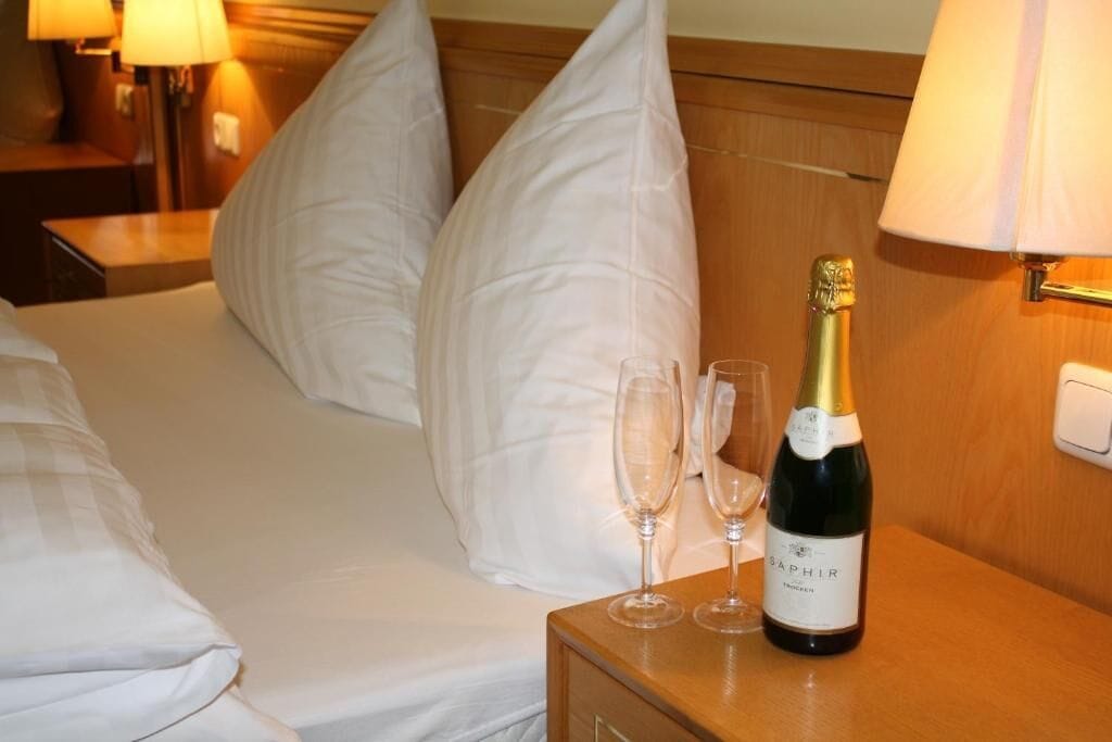 A hotel room with a bed, pillows, a nightstand with a bottle of wine and two glasses.