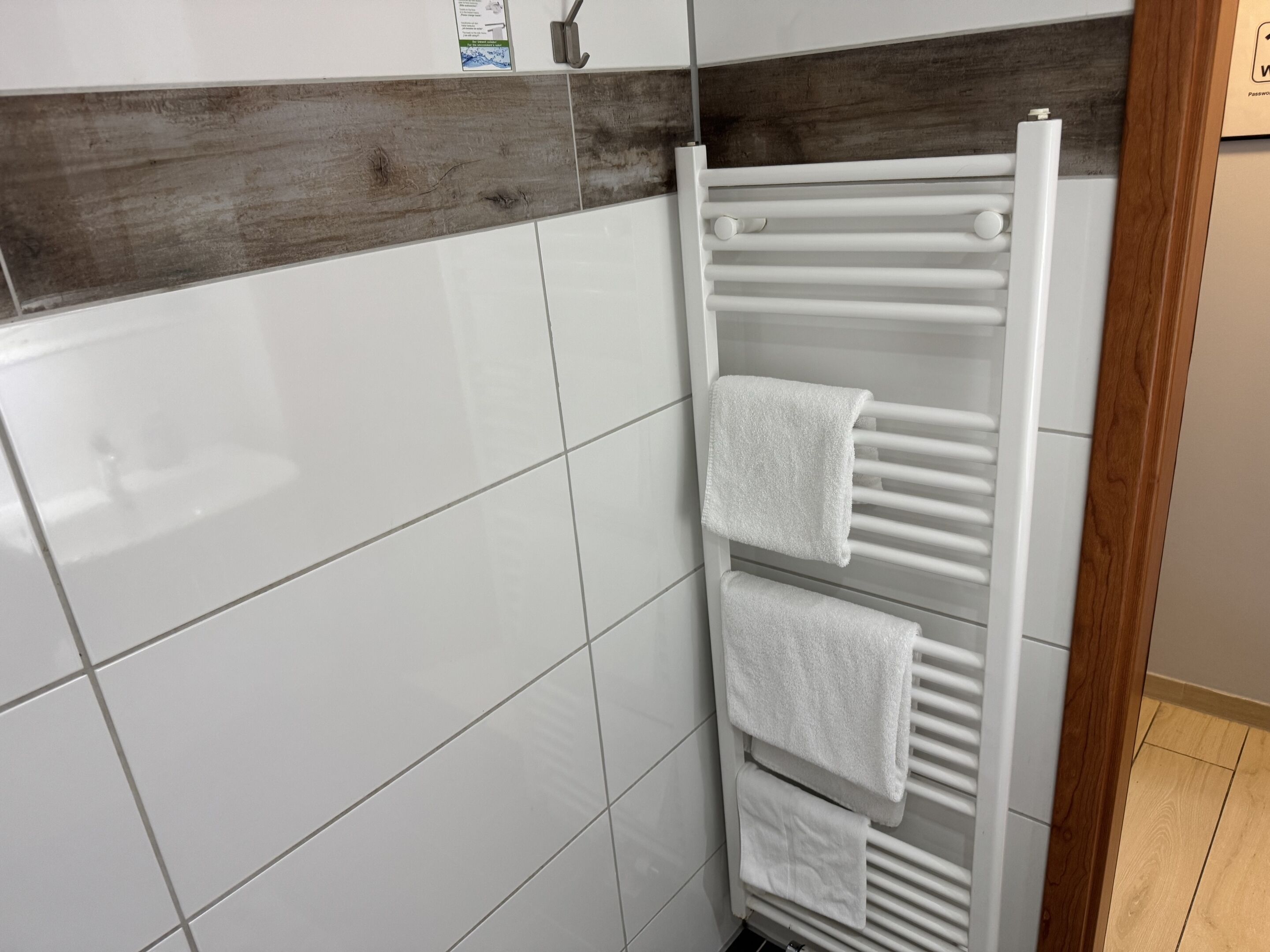 Une salle de bain avec des murs recouverts de carreaux blancs, une bande décorative en bois et un porte-serviettes blanc avec trois serviettes.