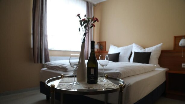 A hotel room with a bed, a bedside table with a bottle and glasses, and a window with curtains. - Hotel Mykonos (Eschweiler)