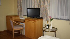 A hotel room with a wooden desk, a television, a small table with a wine bottle and glass, and a chair. - Hotel Mykonos (Eschweiler)