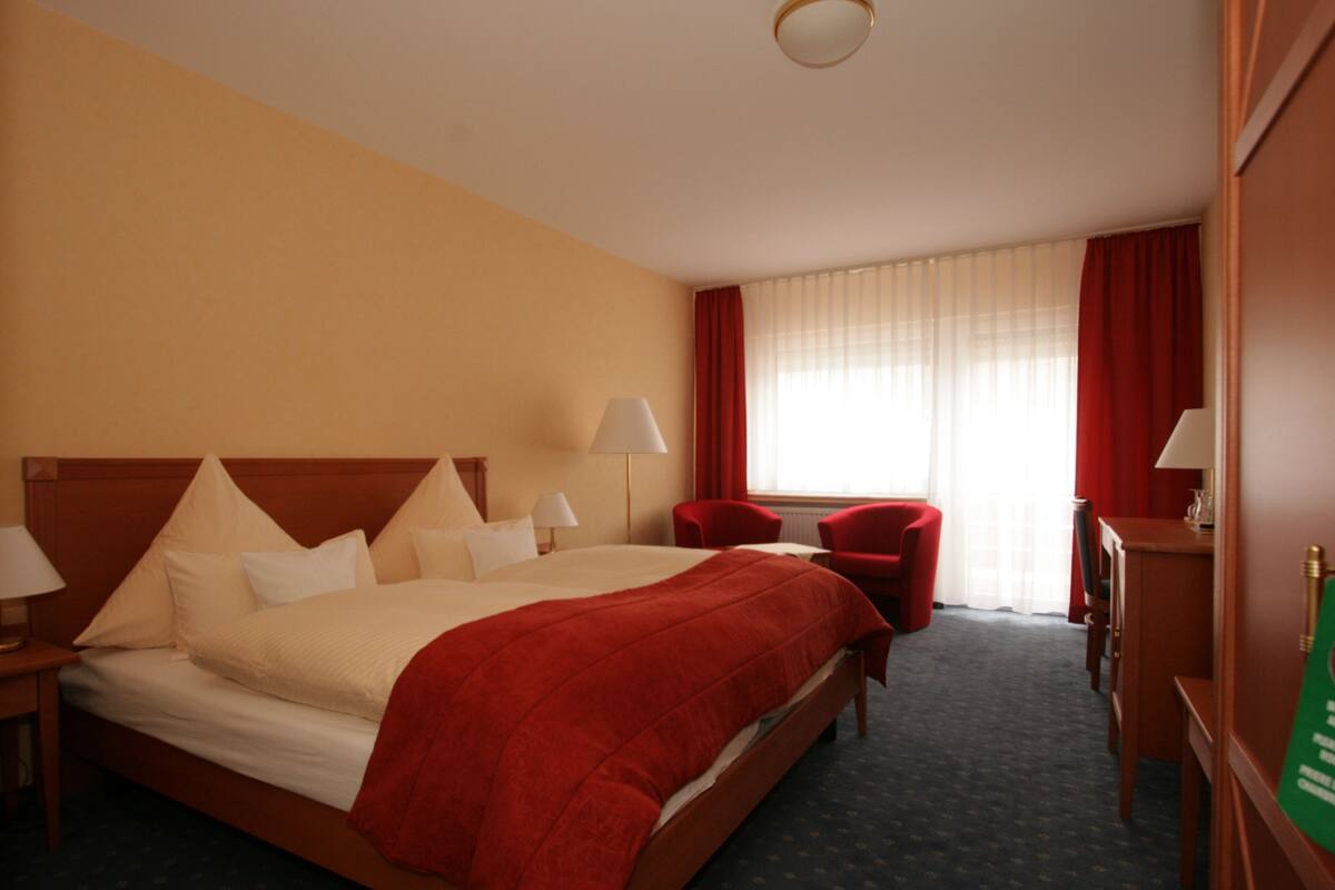 Ein Hotelzimmer mit einem großen Bett, zwei Nachttischen mit Lampen, einem roten Sessel, einem Schreibtisch und einem Fenster mit Vorhängen.