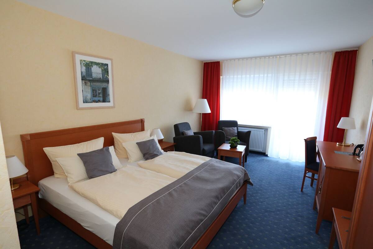 Ein Hotelzimmer mit einem großen Bett, einem Schreibtisch, einem Stuhl, einem kleinen Tisch und einem Fenster mit Vorhängen.